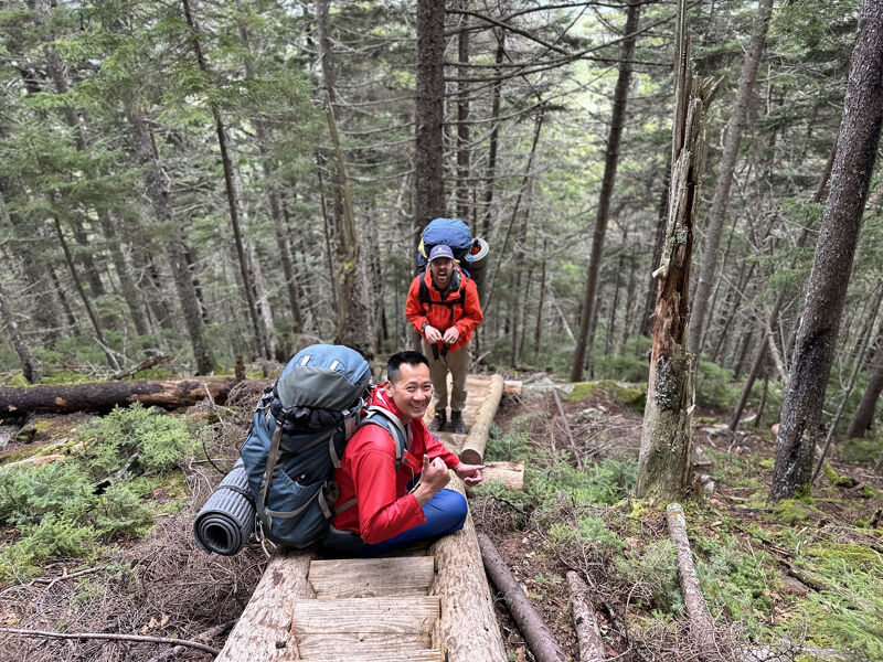The image shows two hikers on a trail in a dense forest. The hiker in the foreground is sitting on a wooden structure, wearing a red jacket and a large backpack. The second hiker is further down the trail, also with a backpack and wearing a red jacket. The trail is surrounded by tall trees and lush greenery, suggesting a wilderness environment.